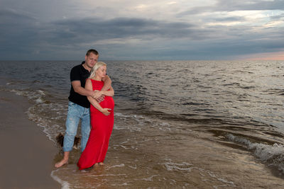 Couple standing at beach against cloudy sky during sunset