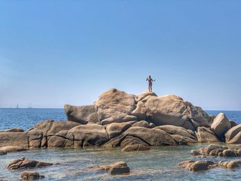 Man standing on rock by sea against clear blue sky