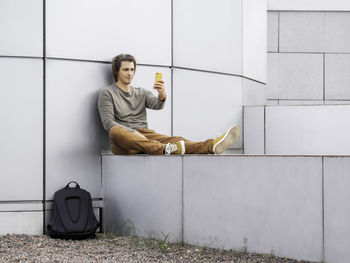 Man is making selfie on smartphone while sitting on wall. student with rucksack in casual clothes. 