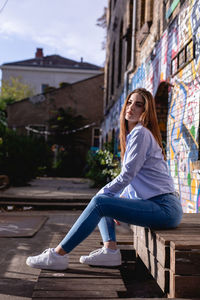 Portrait of confident young woman sitting on wood against wall in city