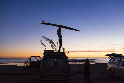 Side view of man carrying surfboard while standing on mini van during vacation with woman on san onofre state beach