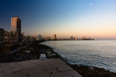 City buildings by sea against clear sky at sunset