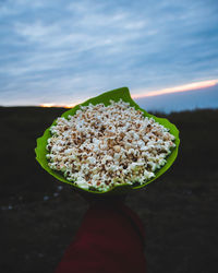 Cropped hand holding popcorn against cloudy sky during sunset