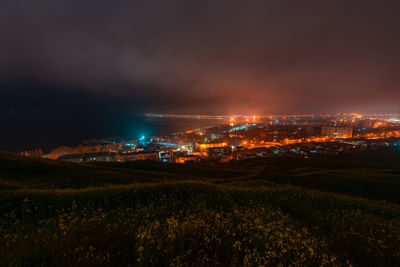 Illuminated field by buildings against sky at night