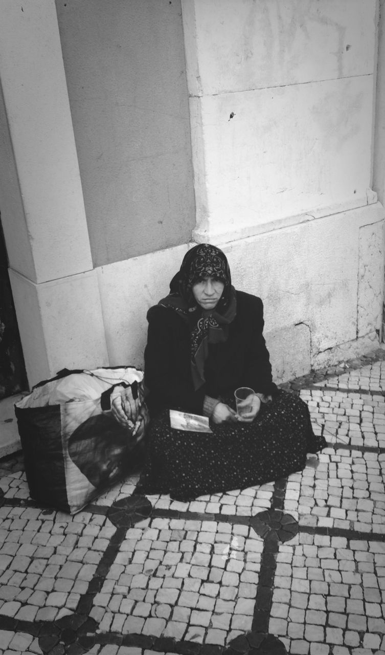 sitting, full length, lifestyles, cobblestone, relaxation, leisure activity, looking at camera, portrait, street, casual clothing, sidewalk, paving stone, young women, day, outdoors, young adult, tiled floor, brick wall