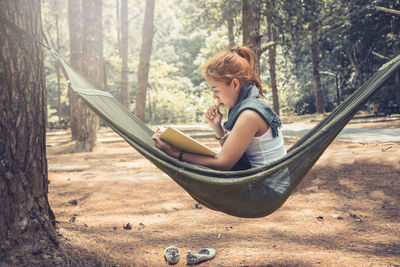 Side view of woman reading book while sitting on hammock in forest