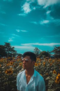 Young man wearing sunglasses looking away while standing on sunflower field