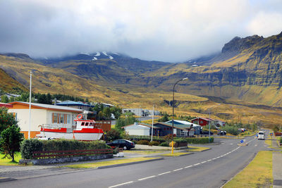 Road by buildings and mountains against sky