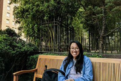 Portrait of smiling woman sitting on bench against trees at park