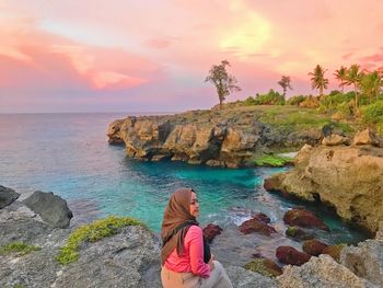 Woman on rock by sea against sky during sunset