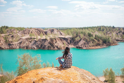 Rear view of woman sitting on rock by lake against sky