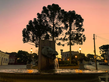 Silhouette tree by illuminated city against orange sky