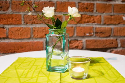 Close-up of tea light candle with flower vase on table against brick wall