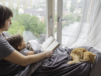 Father reads book to his son. family time on window sill with ginger cat. dad, toddler boy at home.