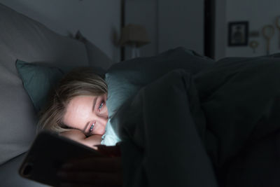 Sleepy tired woman lying in bed under the blanket using mobile phone at night. insomnia. addiction.