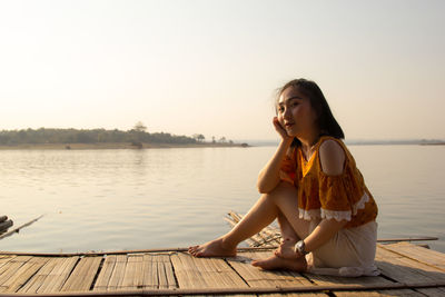 Portrait of woman sitting on wooden raft in lake against clear sky during sunset