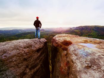 Man in jeans black outdoor sweatshirt and red cap stay at the end of cracked rocky cliff. 