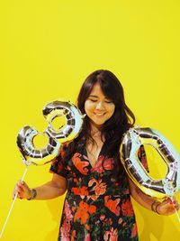 Smiling woman holding helium balloons while standing against yellow background