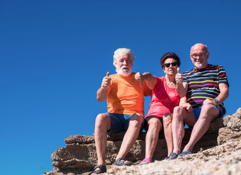 Senior friends gesturing while sitting on rock against clear blue sky