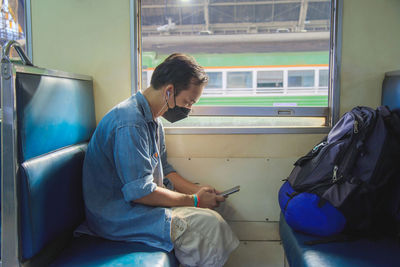 Asian man wearing casual wear and face mask sitting and using a cell phone with earphones on a train 