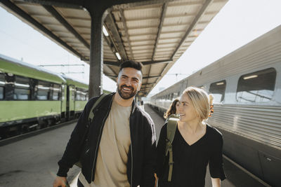 Smiling man walking with woman near train at railroad station