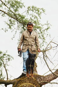 Portrait of young man standing by tree against sky