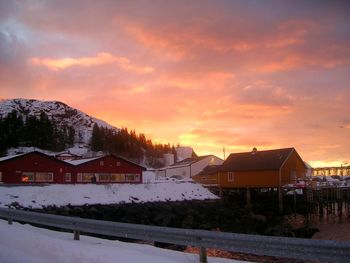 Snow covered houses by buildings against sky during sunset