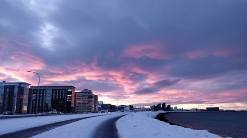 Road amidst snow covered buildings against sky during sunset