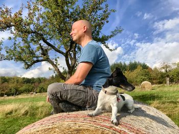 Man with dog sitting on hay bale against sky