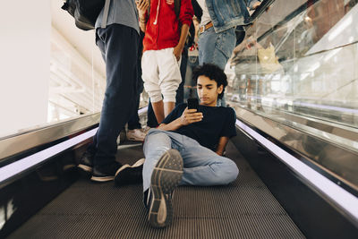 Teenage boy using mobile phone while sitting by friends on moving walkway in shopping mall