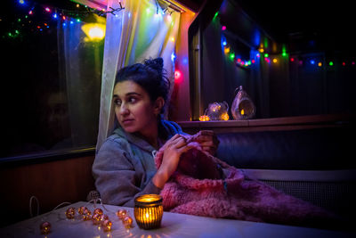 Young woman knitting sweater while sitting in illuminated caravan