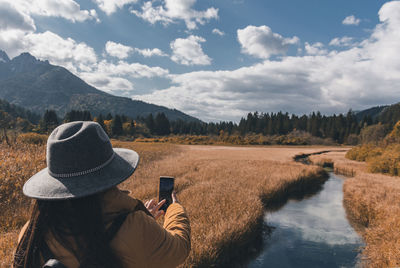 Rear view of woman taking photo of creek flowing through dry grassland