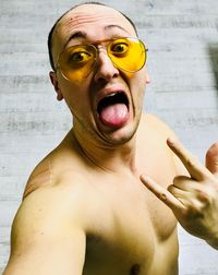 Portrait of shirtless man making a face while gesturing horn sign against wall