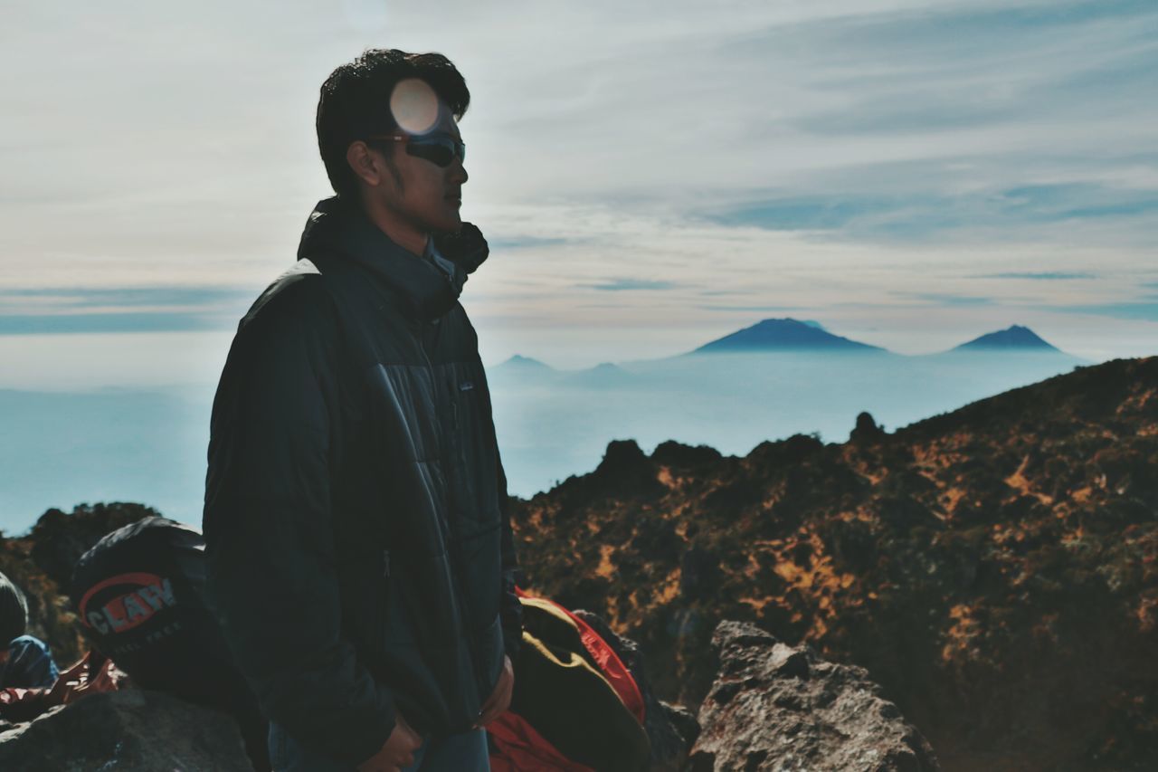 mountain, sunglasses, real people, leisure activity, sky, nature, standing, one person, adventure, outdoors, cloud - sky, young adult, young men, vacations, scenics, day, lifestyles, men, landscape, beauty in nature, young women, sea, water, adult, people