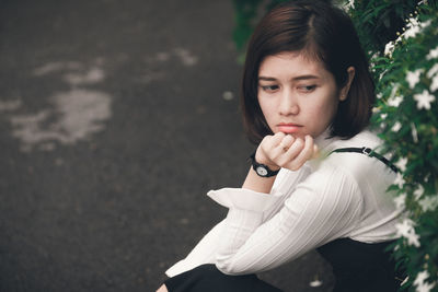 Beautiful young woman looking away while sitting on footpath against plant