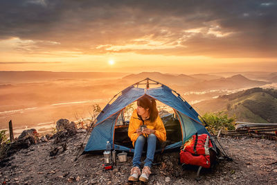 Woman sitting in tent on mountain against sky during sunset