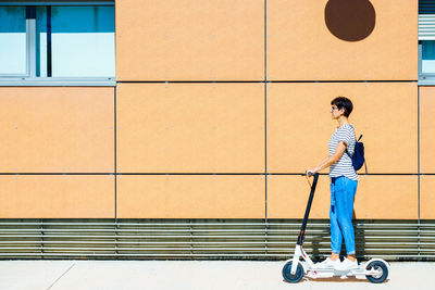 Side view of woman standing on electric push scooter against building
