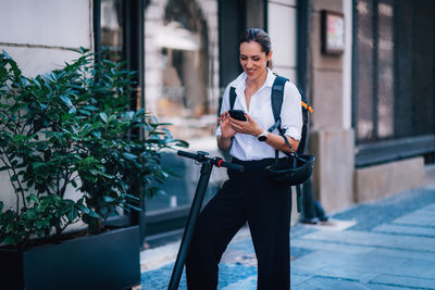 Smiling woman using mobile phone while standing with push scooter in city