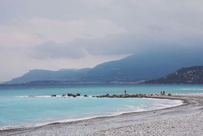 View of sea with rocks by mountains against cloudy sky