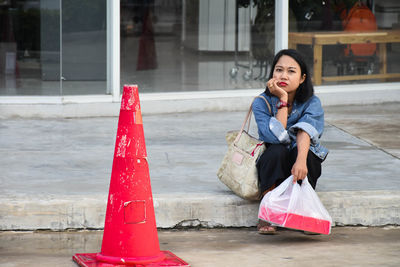 Portrait of woman holding plastic bag while sitting by red traffic cone on road