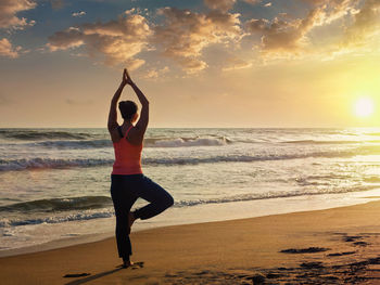 Full length of man with arms raised on beach against sky during sunset