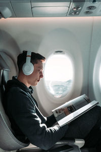 Side view of man with headphones reading magazine while sitting in airplane