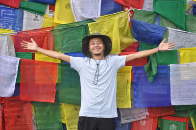 A happy asian male tourist looking at camera with spreading hands against tibetan prayer flags