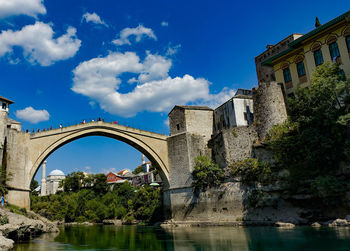 Arch bridge over river by buildings against blue sky