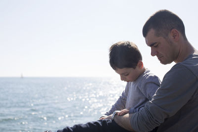 Father and son sitting by sea against clear sky