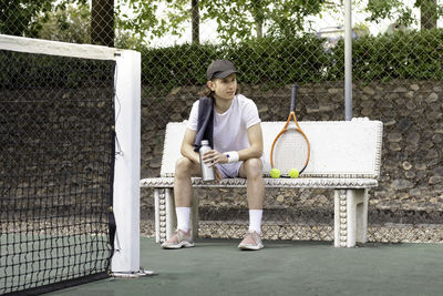 Tired tennis player man looking sideways sitting on a bench on a tennis court with cap on and towel