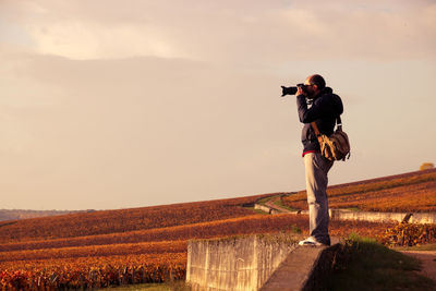 Side view of man photographing on retaining wall against landscape