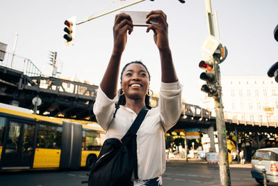 Smiling young woman photographing through mobile phone while standing on street in city