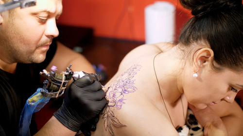 In a tattoo salon , a specialist is doing a tattoo on woman's back, a floral ornament. a man works