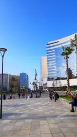 People on footpath by buildings against clear blue sky